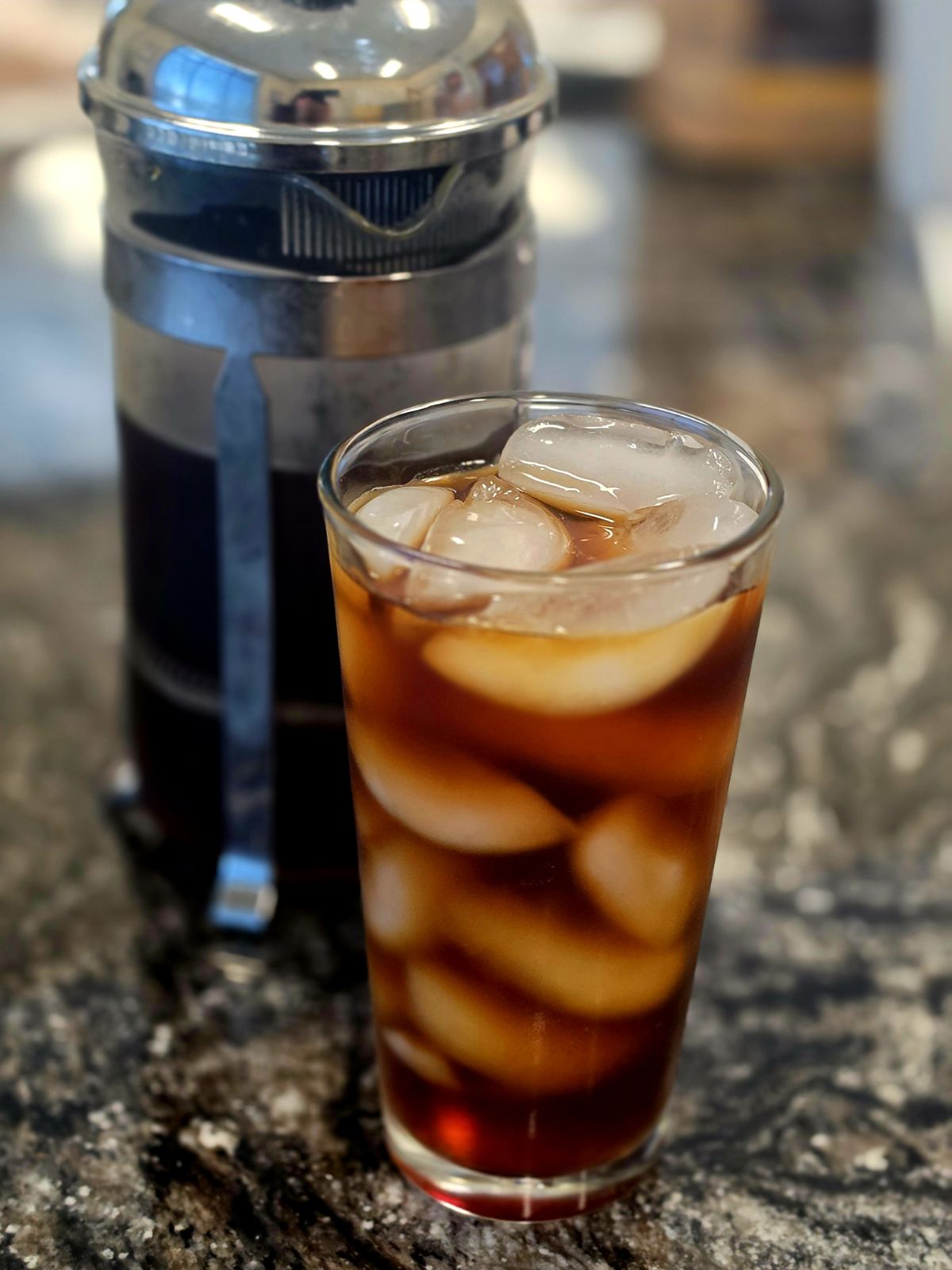 A clear glass tumbler filled with iced coffee and large ice cubes, set on a dark speckled countertop with a blurred metal coffee maker behind it.