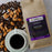 A kraft paper bag of CoffeeAM Almond Coffee Whole Bean lies on a light wood surface, surrounded by scattered coffee beans and almonds, with a white cup of black coffee in the foreground.