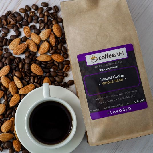 A kraft paper bag of CoffeeAM Almond Coffee Whole Bean lies on a light wood surface, surrounded by scattered coffee beans and almonds, with a white cup of black coffee in the foreground.