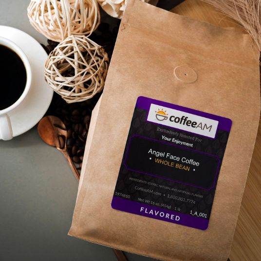 A brown kraft-paper coffee bag labeled "coffeeAM Angel Face Coffee, WHOLE BEAN, FLAVORED" sits on a grey tabletop on top of a light brown cutting board. Positioned nearby are a white ceramic coffee cup on a saucer, scattered whole coffee beans, gold spoon, and decorative woven rattan spheres.