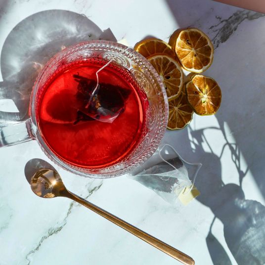 Glass of red tea with a tea bag, dried lemon slices, and a gold spoon on a marble surface.
