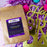 A brown kraft-paper coffee bag labeled "coffeeAM Birthday Cake Coffee, WHOLE BEAN, FLAVORED" lies on a backdrop of shiny purple tinsel, colorful pom-poms, and gold decorative lettering.