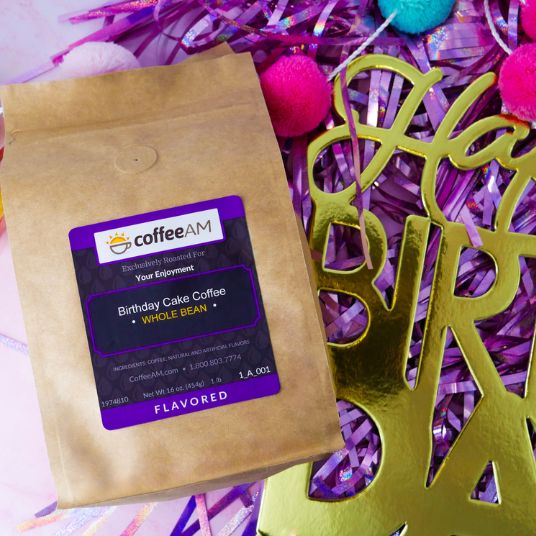 A brown kraft-paper coffee bag labeled "coffeeAM Birthday Cake Coffee, WHOLE BEAN, FLAVORED" lies on a backdrop of shiny purple tinsel, colorful pom-poms, and gold decorative lettering.