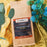 A kraft paper coffee bag labeled coffeeAM Brazil Moreninha Formosa drip grind, placed on shiny gold fabric with decorative green leaves and blue flowers around it.