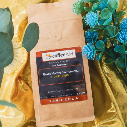 A kraft paper coffee bag labeled coffeeAM Brazil Moreninha Formosa drip grind, placed on shiny gold fabric with decorative green leaves and blue flowers around it.
