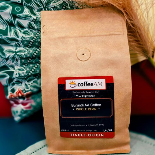 A bag of coffeeAM Burundi AA Coffee Whole Bean, single-origin, in a brown kraft paper pouch with a black and red label, held in front of a patterned green and white fabric background.