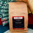 A bag of coffeeAM Burundi AA Coffee Whole Bean, single-origin, in a brown kraft paper pouch with a black and red label, held in front of a patterned green and white fabric background.