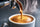 A close-up of espresso being poured from a metal portafilter into a white ceramic cup, with visible steam rising against a dark gray background.