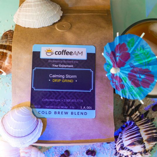 A bag of CoffeeAM Calming Storm drip grind cold brew blend, styled with seashells and a blue floral cocktail umbrella on a turquoise surface.
