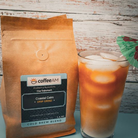 A brown stand-up coffee bag labeled coffeeAM Coastal Calm drip grind Cold Brew Blend next to a tall glass of iced coffee with ice and a green cocktail umbrella, set against a rustic whitewashed wood background.
