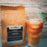 A brown stand-up coffee bag labeled coffeeAM Coastal Calm drip grind Cold Brew Blend next to a tall glass of iced coffee with ice and a green cocktail umbrella, set against a rustic whitewashed wood background.