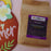 A brown kraft paper bag of coffeeAM Coconut Cream Coffee, Whole Bean, with a purple and black label, shown next to a decorative wooden sign on a pink background.