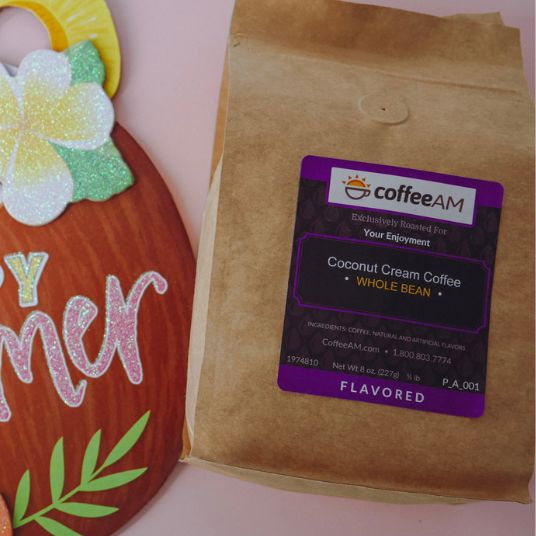 A brown kraft paper bag of coffeeAM Coconut Cream Coffee, Whole Bean, with a purple and black label, shown next to a decorative wooden sign on a pink background.