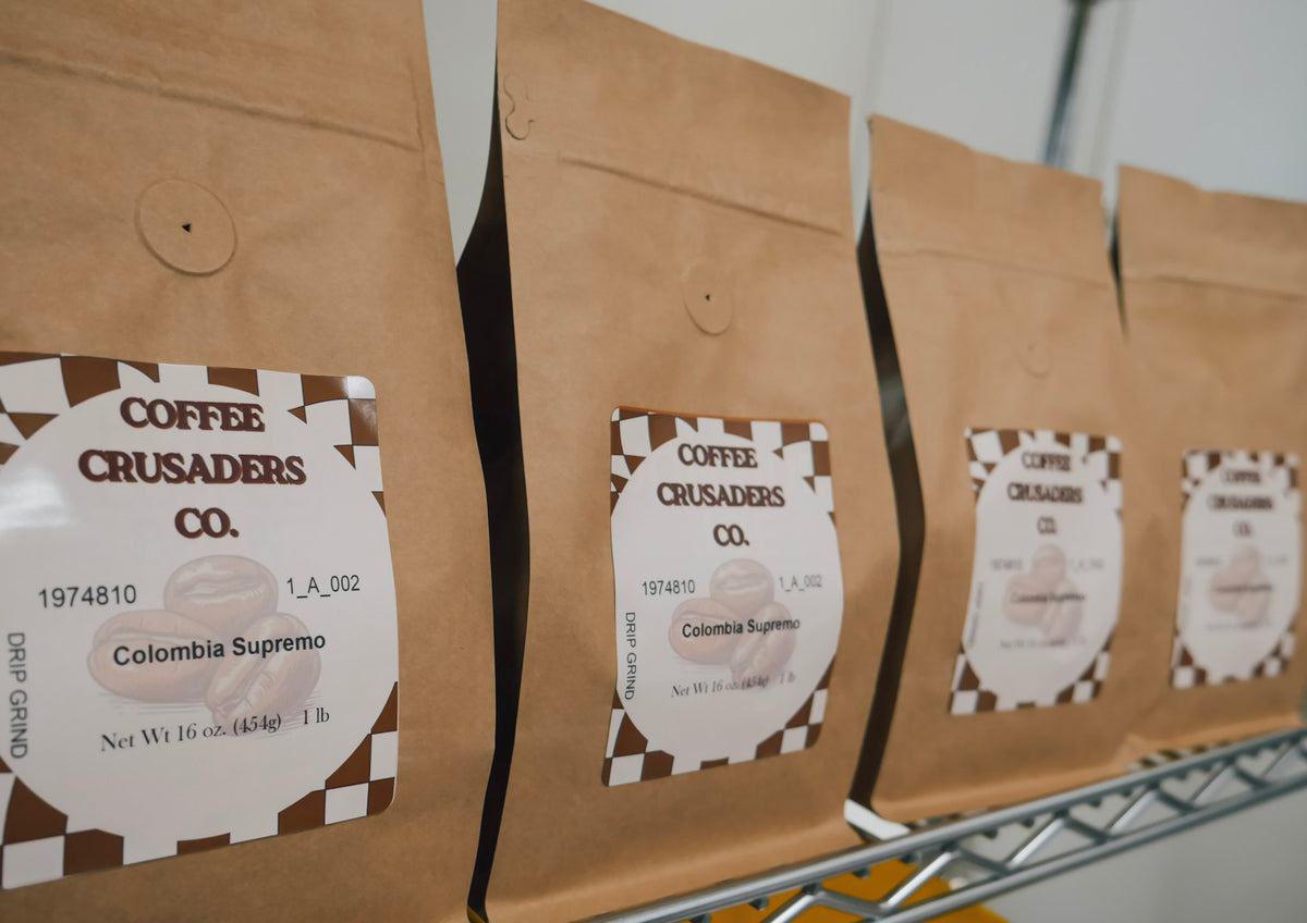 Several brown paper coffee bags on a metal shelf, each with a white label reading Coffee Crusaders Co. and Colombia Supremo.