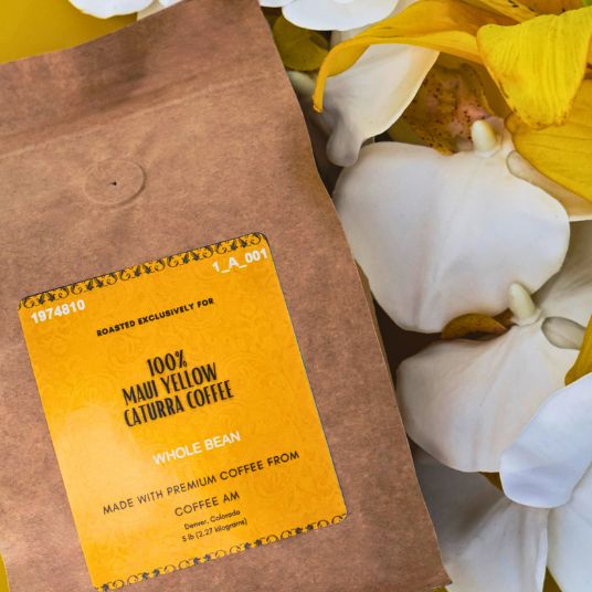 A kraft paper coffee bag with a yellow label reading '100% Marigold Catumbia Coffee' and 'LA-941', styled on a bright yellow background with white and yellow flowers.