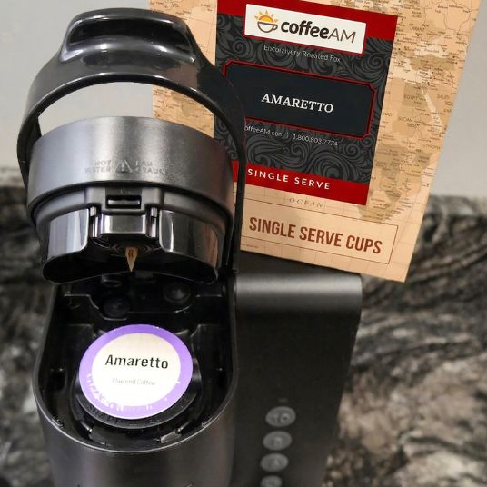 Box of Coffee AM Amaretto single serve cups with a glass of iced coffee in the background.