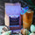 A bag of CoffeeAM Decaf Coastal Calm cold brew coffee stands next to an iced coffee drink with a paper cocktail umbrella, styled with seashells and coffee beans on a marble surface.
