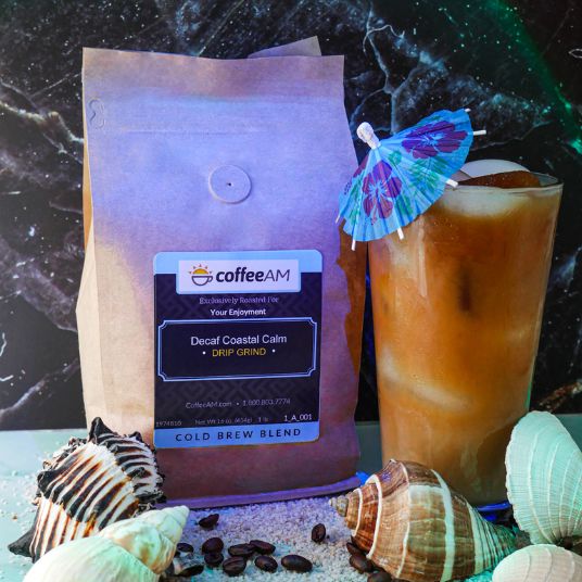 A bag of CoffeeAM Decaf Coastal Calm cold brew coffee stands next to an iced coffee drink with a paper cocktail umbrella, styled with seashells and coffee beans on a marble surface.
