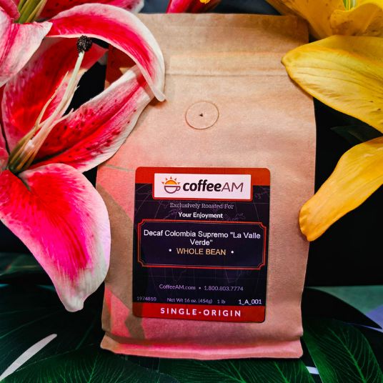 A stand-up kraft paper coffee bag labeled CoffeeAM Decaf Colombia Supremo "La Valle Verde" Whole Bean, surrounded by large pink and yellow flowers and green leaves.