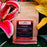 A stand-up kraft paper coffee bag labeled CoffeeAM Decaf Colombia Supremo "La Valle Verde" Whole Bean, surrounded by large pink and yellow flowers and green leaves.