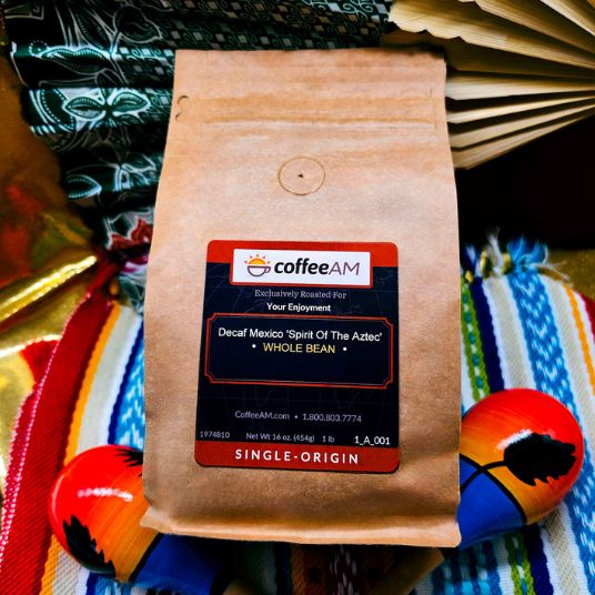 A bag of CoffeeAM Decaf Mexico 'Spirit Of The Aztecs' whole bean coffee, placed on a colorful Mexican serape blanket with decorative maracas and a fan in the background.