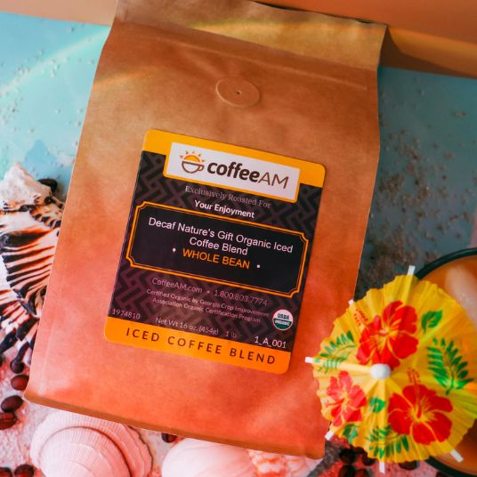A bag of Coffee AM Decaf Nature's Gift Organic Iced Coffee Blend Whole Bean with a yellow and black label, styled on a light blue surface with seashells and a yellow tropical cocktail umbrella, in a glass of iced coffee