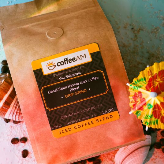 A bag of coffeeAM Decaf Spirit Revive Iced Coffee Blend Drip Grind with a black and yellow label, shown at an angle with coffee beans and a colorful paper umbrella on a textured teal background, surrounded with seashells and coffee beans.