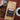A bag of coffeeAM Espresso Prima French Press coffee lies on a dark red surface, surrounded by roasted coffee beans, a metal portafilter, and a cup of black coffee on a saucer. The bag has a brown kraft-paper look with a dark label featuring the coffeeAM brand logo.