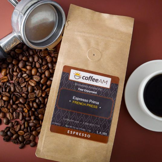 A bag of coffeeAM Espresso Prima French Press coffee lies on a dark red surface, surrounded by roasted coffee beans, a metal portafilter, and a cup of black coffee on a saucer. The bag has a brown kraft-paper look with a dark label featuring the coffeeAM brand logo.