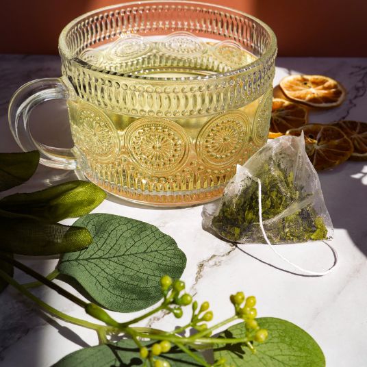 Decorative glass mug with hot liquid, tea bag, and green leaves on a marble surface.