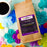 A bag of CoffeeAM Hawaiian Hazelnut Coffee French Press flavor, styled in a flat-lay with a white coffee cup of black coffee and colorful artificial flowers on a mint green background.