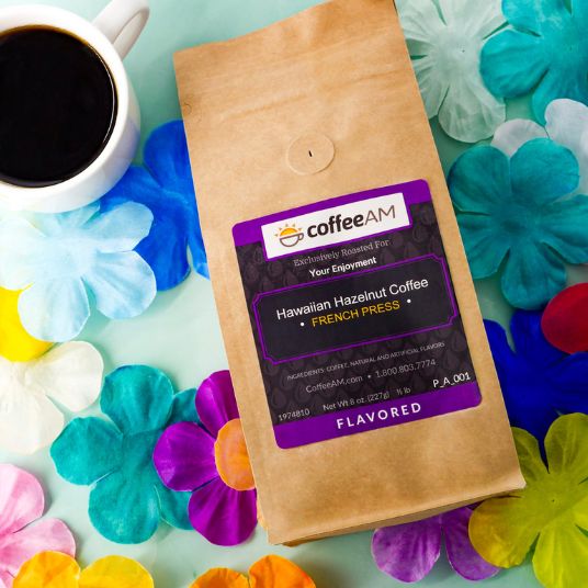 A bag of CoffeeAM Hawaiian Hazelnut Coffee French Press flavor, styled in a flat-lay with a white coffee cup of black coffee and colorful artificial flowers on a mint green background.