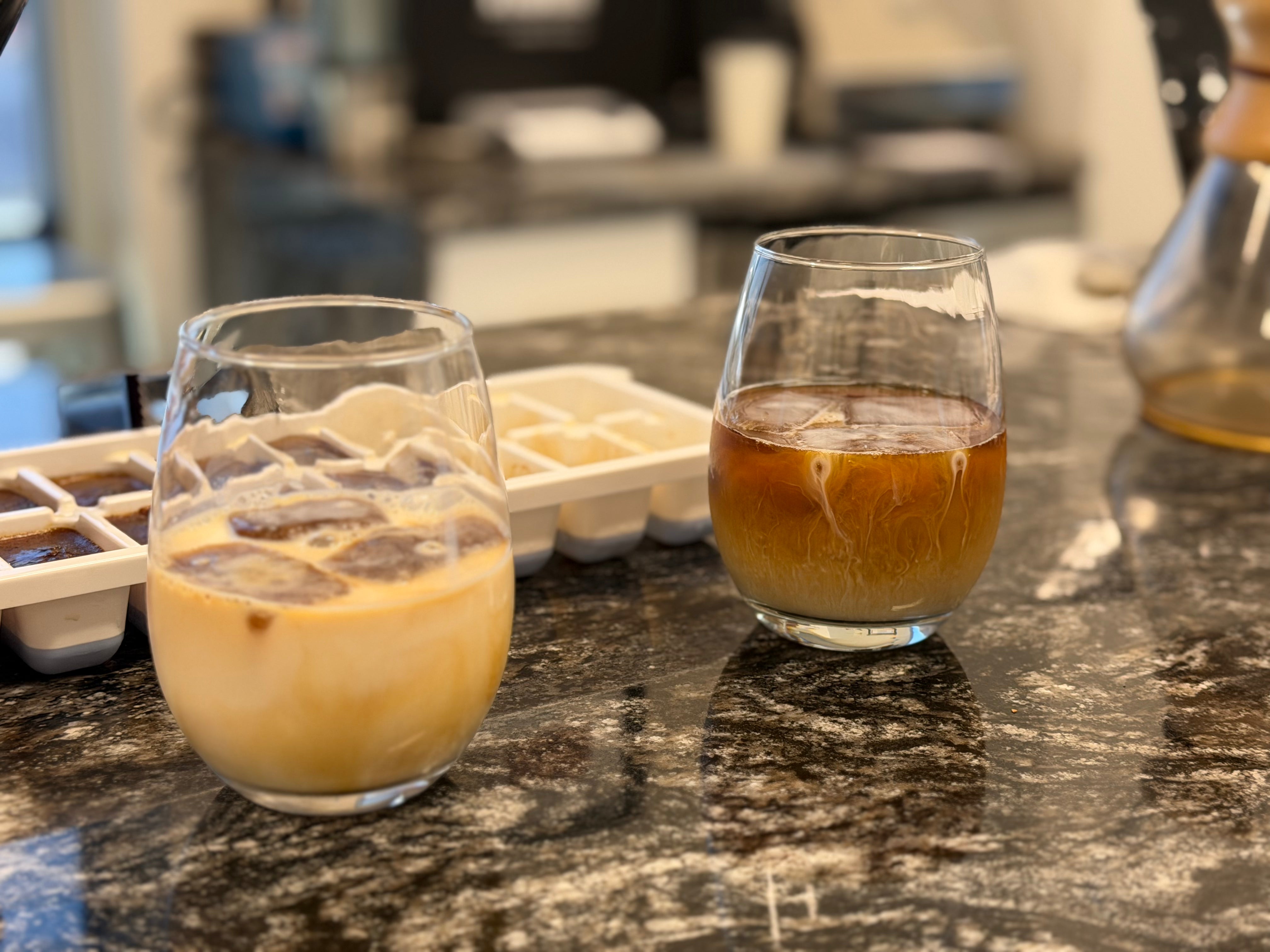 Two clear stemless wine glasses on a dark marble countertop, one filled with iced coffee and cream, and the other with iced coffee and amber liquid. An ice cube tray and a carafe are blurred in the background.