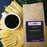 A kraft paper bag of coffeeAM Jamaican Rum Coffee sits next to a white cup of black coffee, surrounded by yellow corn husks and green palm leaves on a dark blue surface.