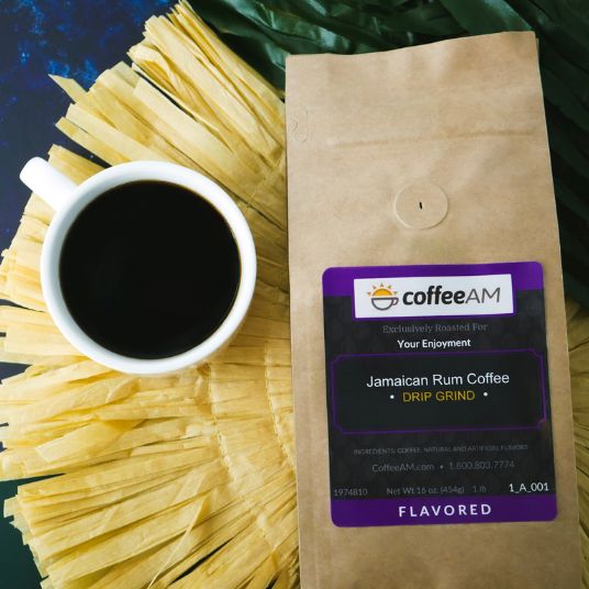 A kraft paper bag of coffeeAM Jamaican Rum Coffee sits next to a white cup of black coffee, surrounded by yellow corn husks and green palm leaves on a dark blue surface.