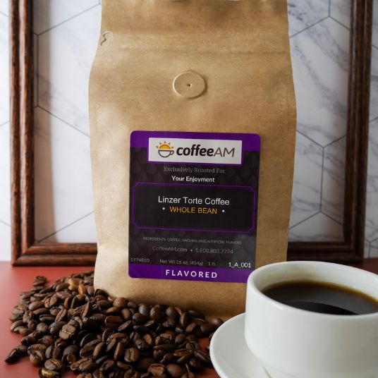 A brown kraft-paper coffee bag labeled "coffeeAM Linzer Torte Coffee, WHOLE BEAN, FLAVORED" stands in front of a framed picture frame on a white marble surface. Whole roasted coffee beans are scattered at the base of the bag on a red cloth, next to a white ceramic mug of black coffee in the foreground.