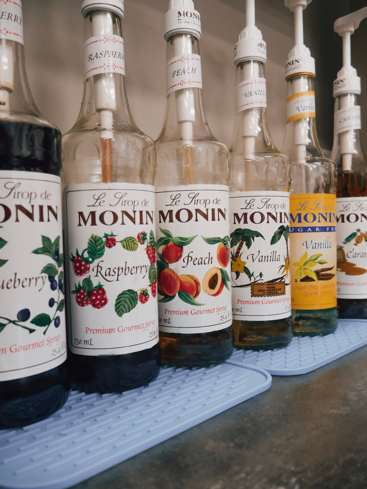 A row of Le Trop de Monin syrup bottles with built-in pumps, including Raspberry, Peach, and Vanilla flavors, lined up on a light blue textured mat.