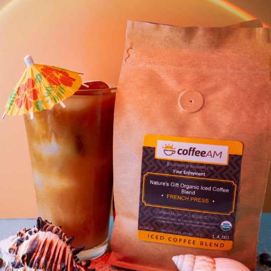A bag of Coffee AM Nature's Gift Organic Iced Coffee Blend French Press with a black and yellow label, next to an iced coffee drink with a tropical paper umbrella, set against a warm orange background, and surrounded by seashells