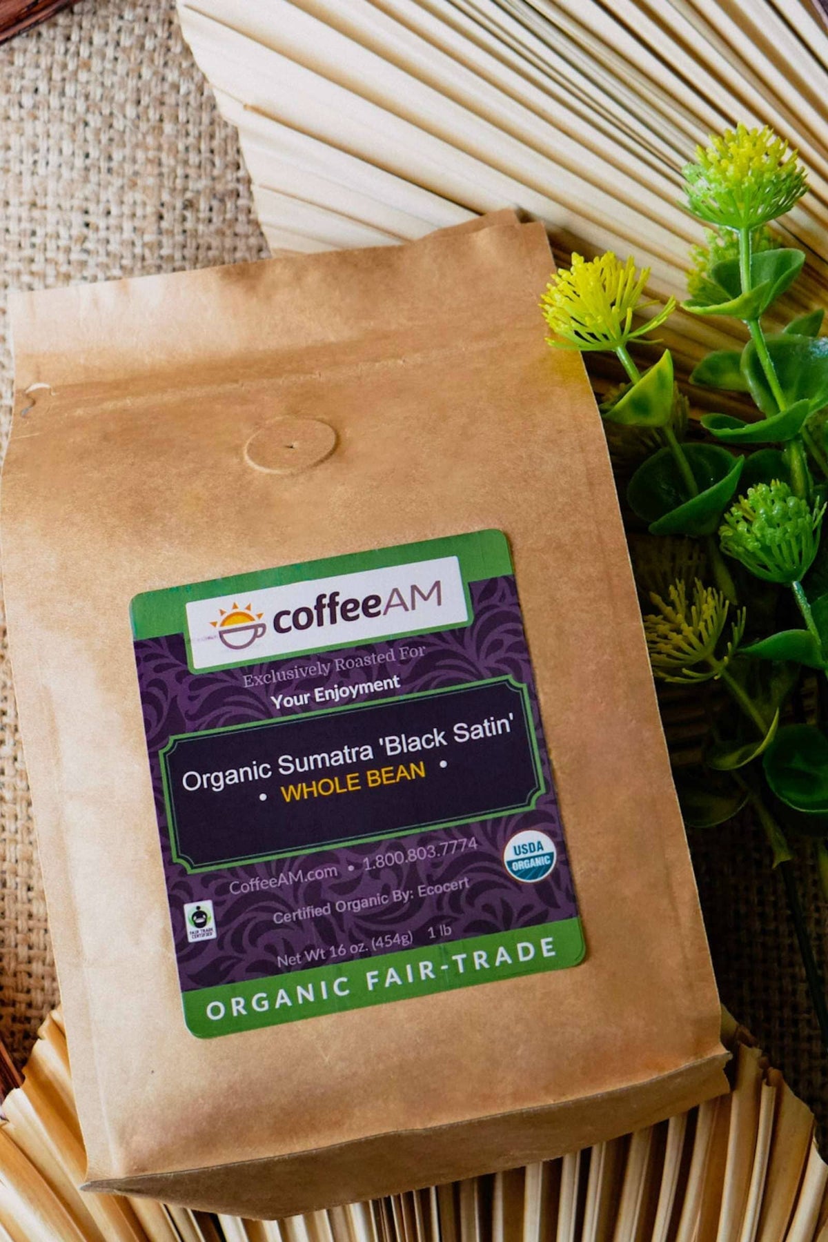 A kraft paper bag of CoffeeAM Organic Sumatra 'Black Satin' Whole Bean coffee, styled against a burlap background with a decorative fan and green foliage with yellow accents. The label displays the CoffeeAM logo, USDA Organic seal, and reads 'ORGANIC FAIR-TRADE'.