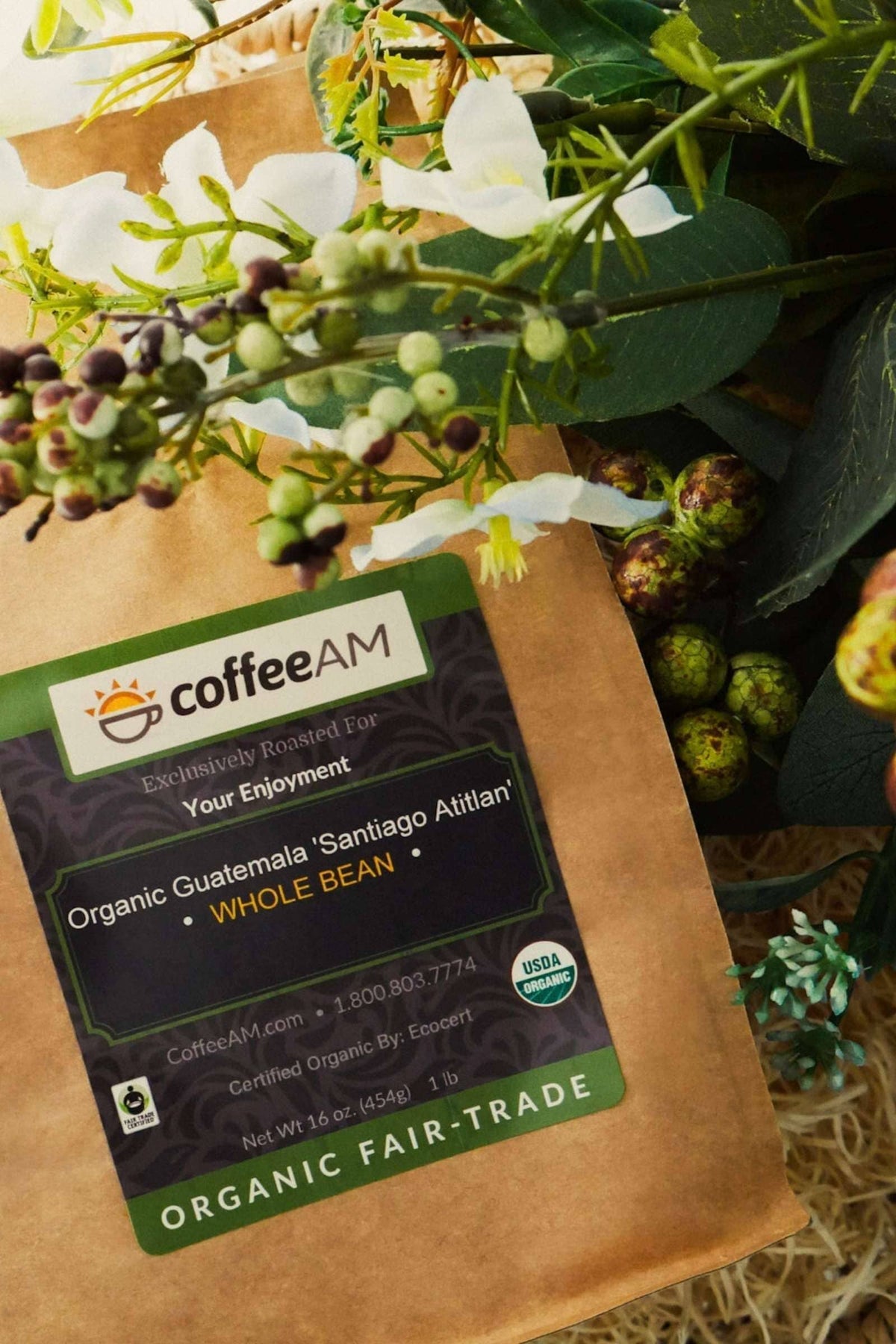 A kraft paper bag of CoffeeAM Organic Guatemala 'Santiago Atitlan' Whole Bean coffee, styled against a background of green foliage, white flowers, and coffee cherries. The label displays the CoffeeAM logo, USDA Organic seal, and reads 'ORGANIC FAIR-TRADE'.