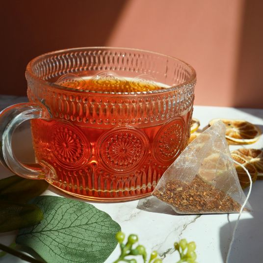 Glass mug with patterned design filled with tea, next to a tea bag and leaves on a light surface.