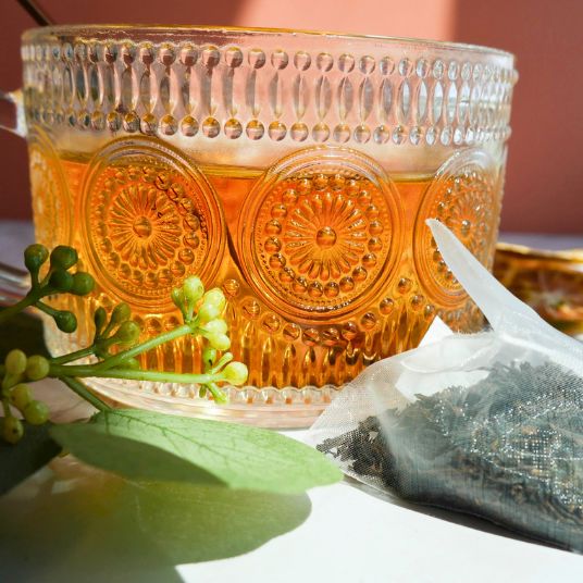 Decorative glass mug with patterned design filled with a warm beverage, next to a tea bag and green leaves on a white surface.