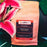 A bag of CoffeeAM Panama Boquete whole bean coffee placed on dark green tropical leaves next to a pink lily flower. The coffee bag is kraft brown with a prominent black and red label.