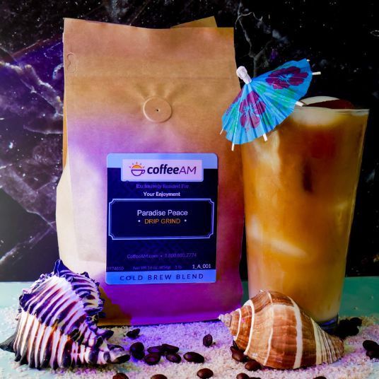 A bag of coffeeAM Paradise Peace cold brew coffee stands next to a tall glass of iced coffee with a colorful paper umbrella, styled with seashells and coffee beans on sand.