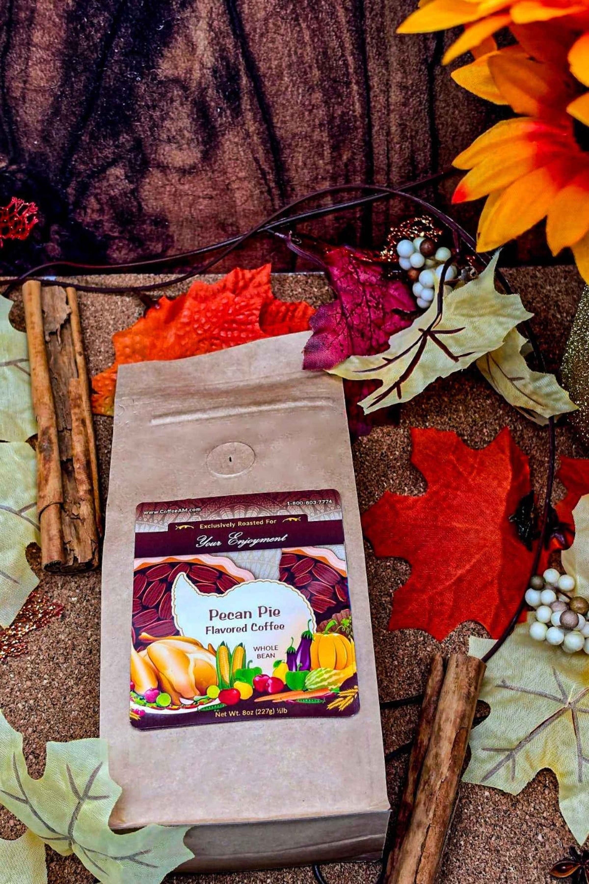 A kraft paper bag of CoffeeAM "Pecan Pie" flavored coffee positioned against an autumn-themed background featuring colorful orange, red, and yellow leaves, cinnamon sticks, and festive holiday decor.