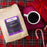A bag of CoffeeAM Peppermint Coffee sits on a red plaid blanket next to a white cup of black coffee on a magenta tray, with a red gift box and candy canes nearby.
