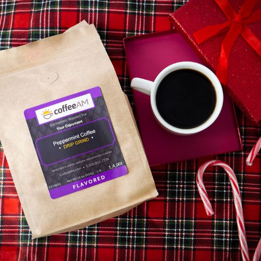 A bag of CoffeeAM Peppermint Coffee sits on a red plaid blanket next to a white cup of black coffee on a magenta tray, with a red gift box and candy canes nearby.