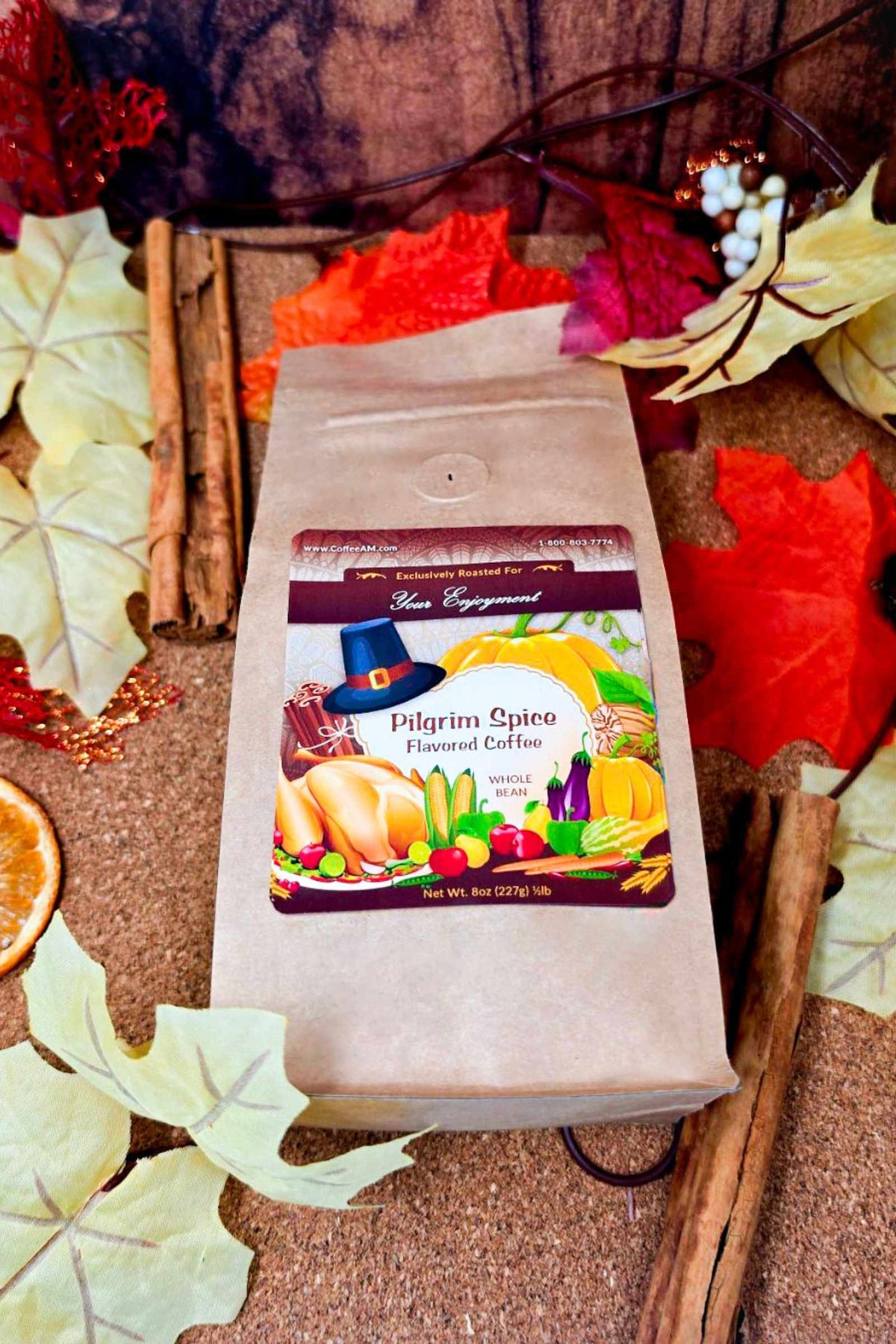 A kraft paper bag of CoffeeAM "Pilgrim Spice" flavored coffee centered against an autumn-themed background featuring colorful red, orange, and yellow leaves, cinnamon sticks, dried orange slices, and white berry accents.