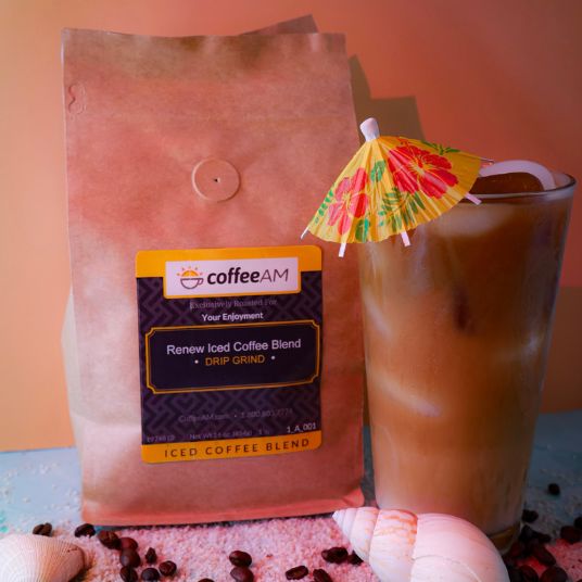 A bag of Coffee AM Renew Iced Coffee Blend next to a glass of iced coffee with a small tropical paper umbrella, styled with seashells on a pink sand with coffee beans and a blue surface against a peach background.