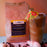 A bag of Coffee AM Renew Iced Coffee Blend next to a glass of iced coffee with a small tropical paper umbrella, styled with seashells on a pink sand with coffee beans and a blue surface against a peach background.